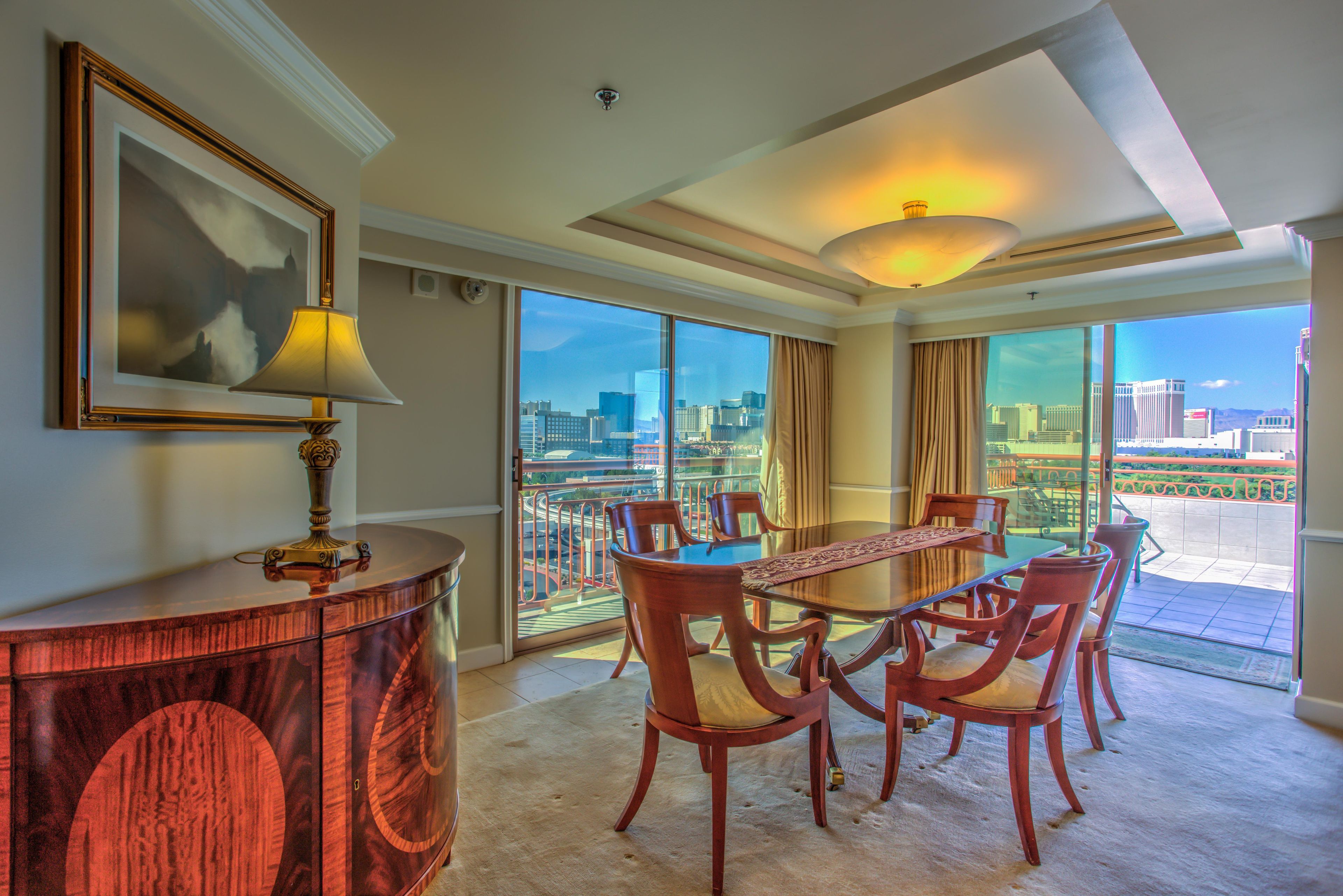 A hotel room with a dining table, chairs, a lamp, a painting, and a large window with a city view.