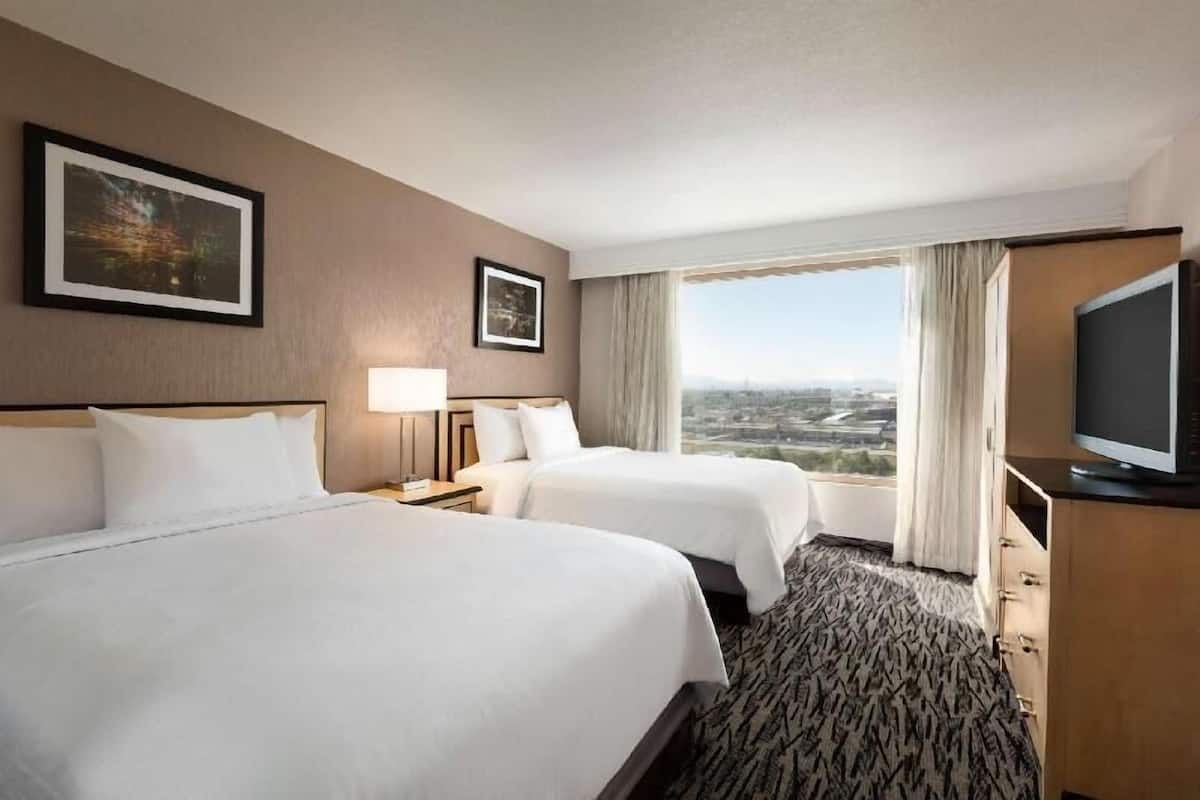 Een hotelkamer met twee bedden, een televisie, een nachtkastje, een lamp en een groot raam met uitzicht op de stad.