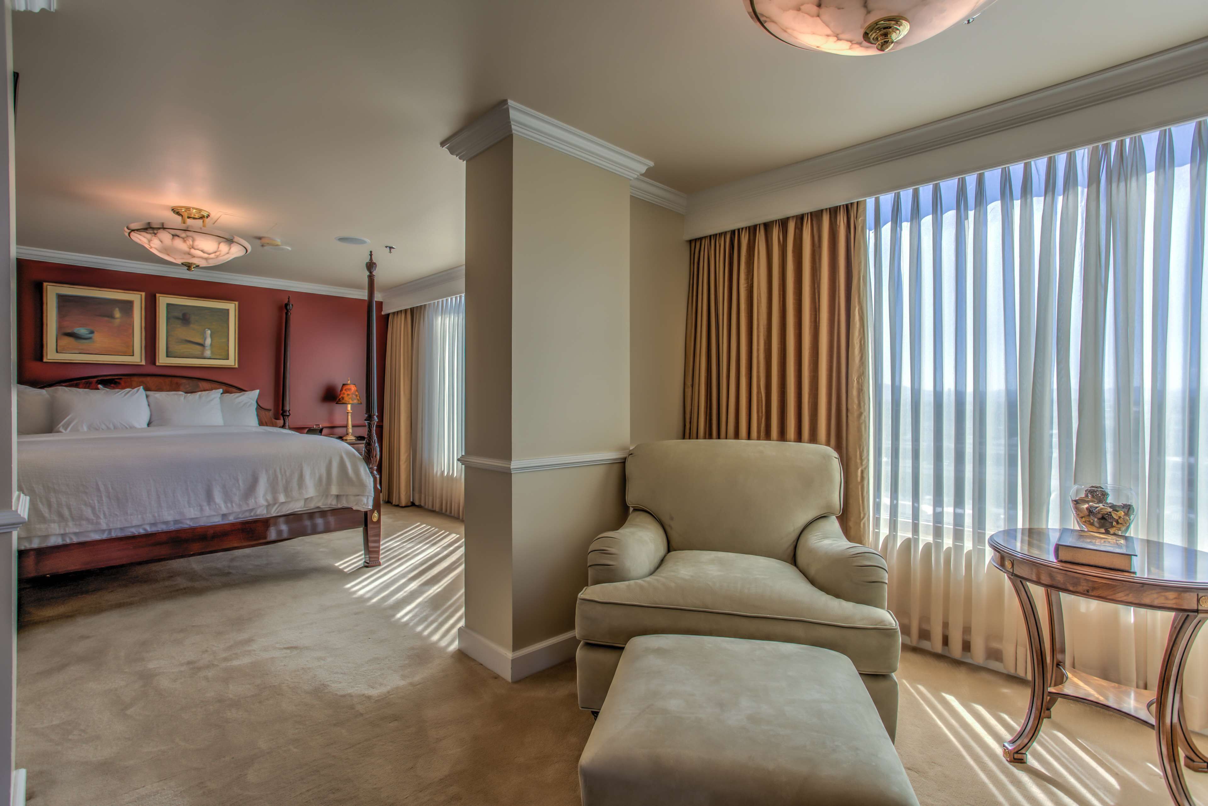 A hotel room with a bed, a chair, a table, and a large window with curtains.