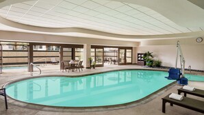 Een binnenzwembad met helder water, omgeven door ligbedden en tafels, en een plafond met een rasterpatroon.