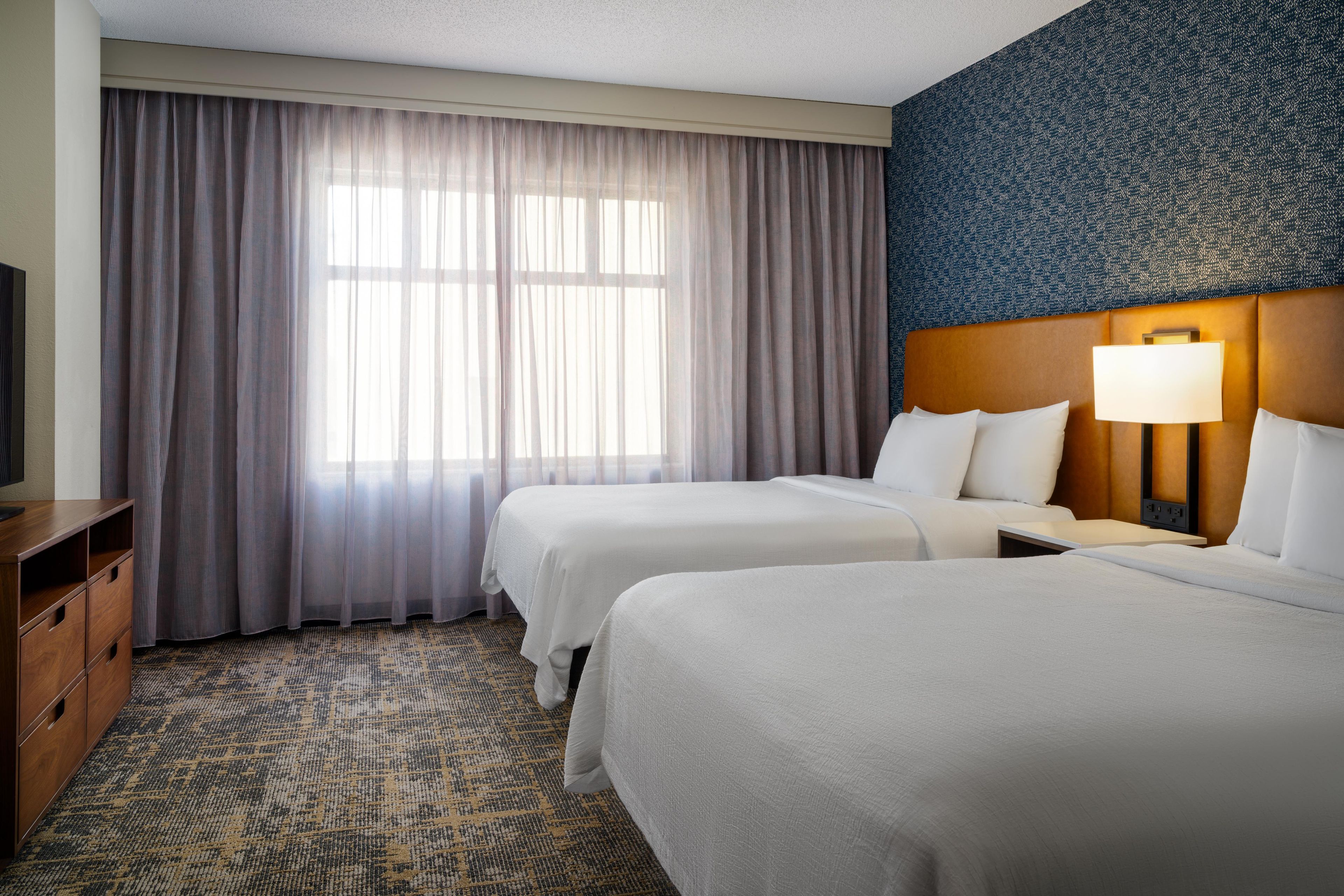 A hotel room with two beds, a television, a desk, and a patterned carpet.