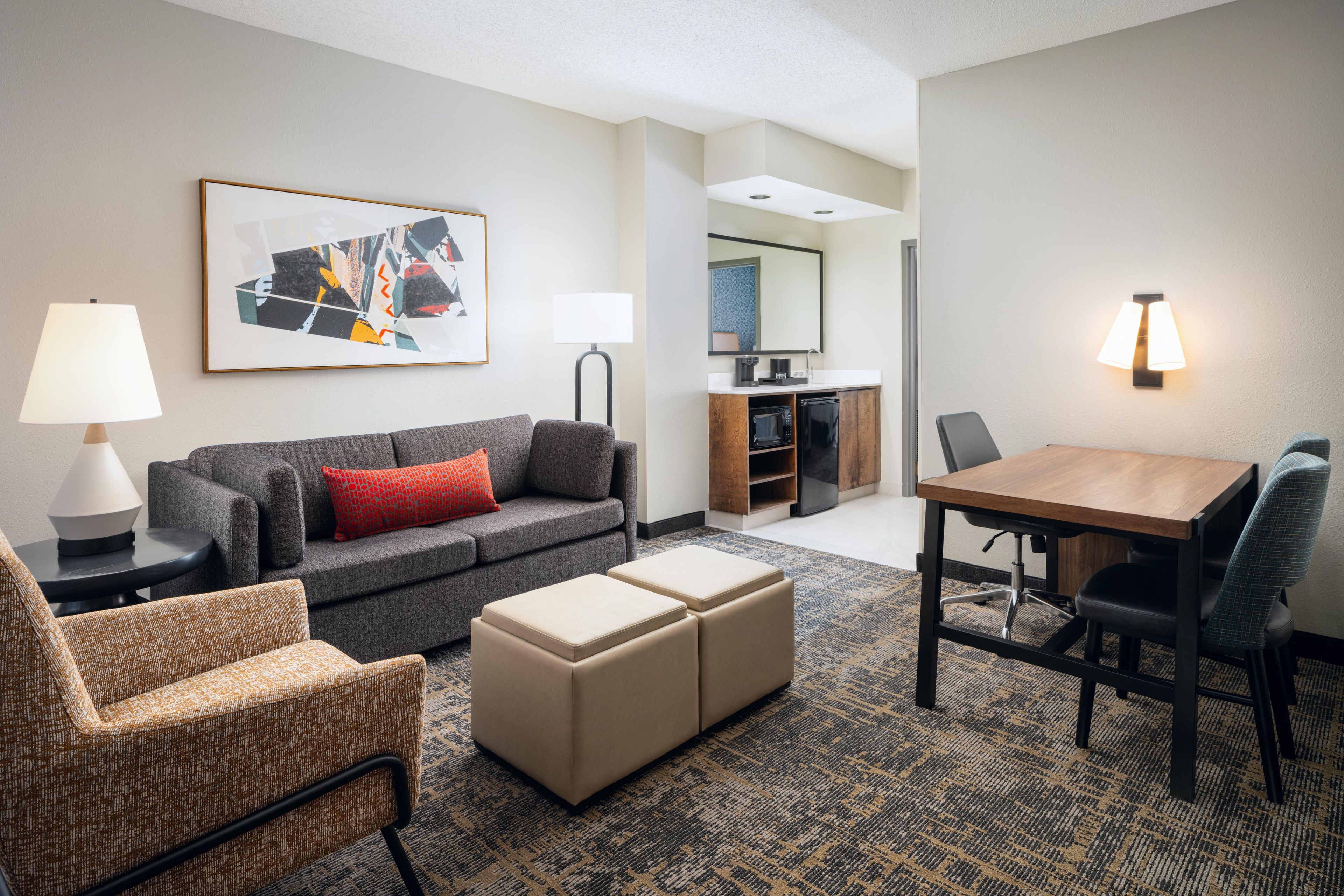 A hotel room with a gray sofa, a small dining table, a kitchenette, and a large window.
