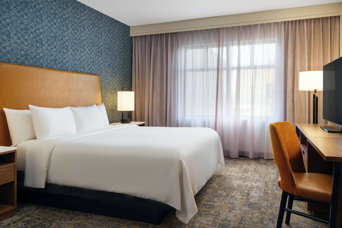 A hotel room with a large bed, a desk with a chair, a TV, and a window with curtains.