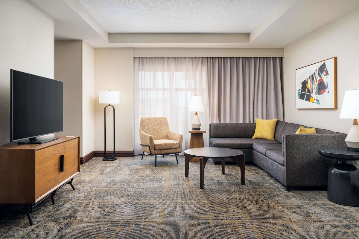 A hotel room with a flat-screen TV, a sofa, a chair, a small round table, and a patterned carpet.
