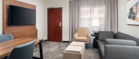 A hotel room with a wooden headboard, a flat-screen TV, a sofa, a chair, a table, and a lamp.
