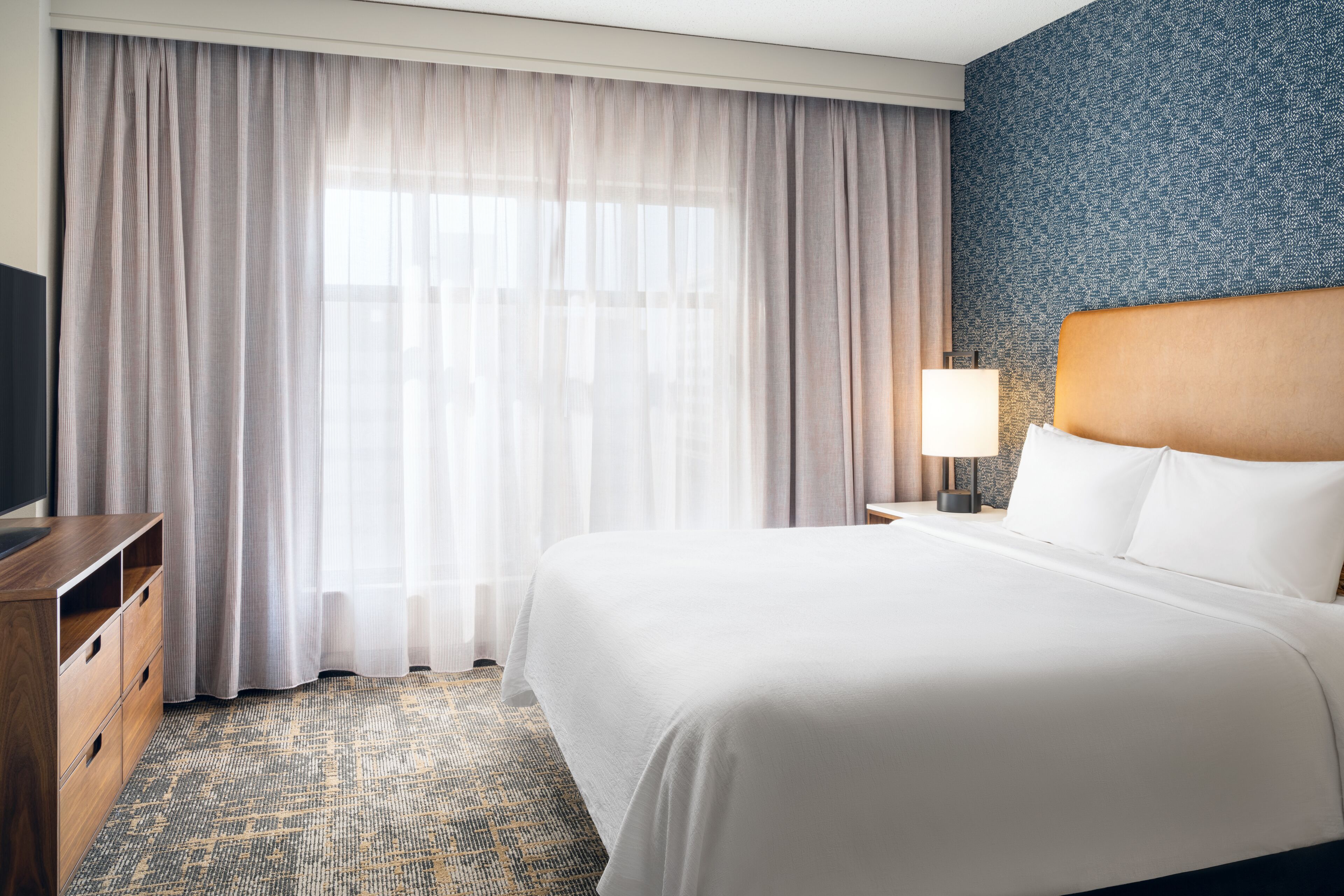 A hotel room with a large bed, a wooden dresser, a nightstand with a lamp, and a window with sheer curtains.