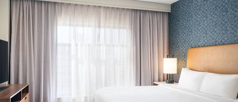 A hotel room with a large bed, a wooden dresser, a nightstand with a lamp, and a window with sheer curtains.