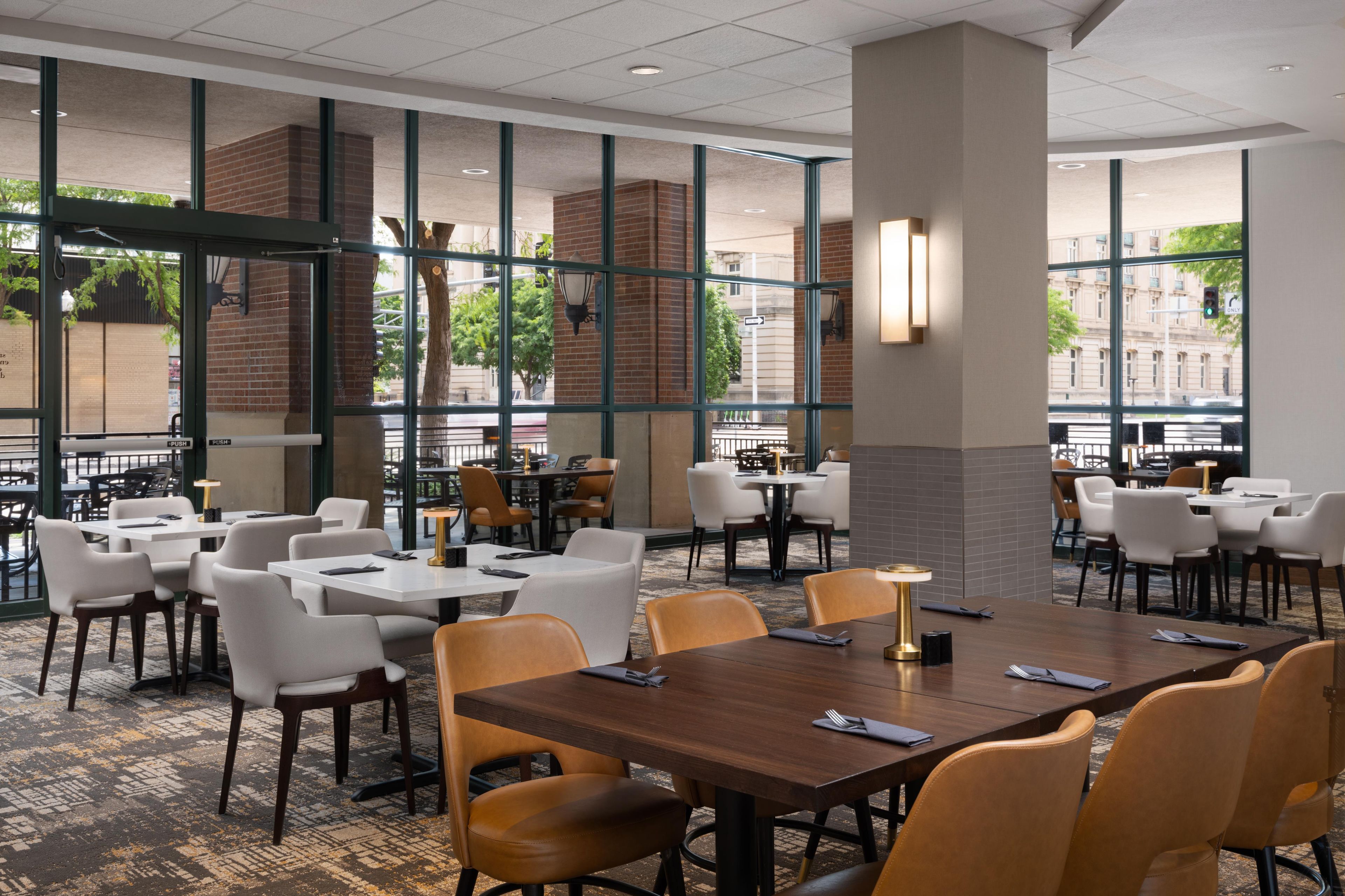A hotel dining area with multiple tables and chairs, large windows, and a view of the street.