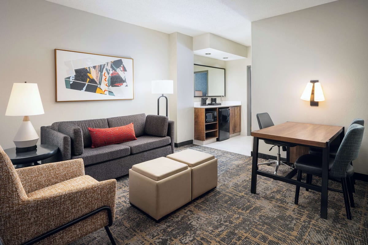 A hotel room with a gray sofa, a small wooden table, a chair, a lamp, and a kitchen area in the background.