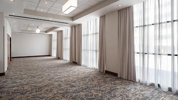A spacious hotel room with a patterned carpet, large windows with curtains, and a ceiling with recessed lighting.