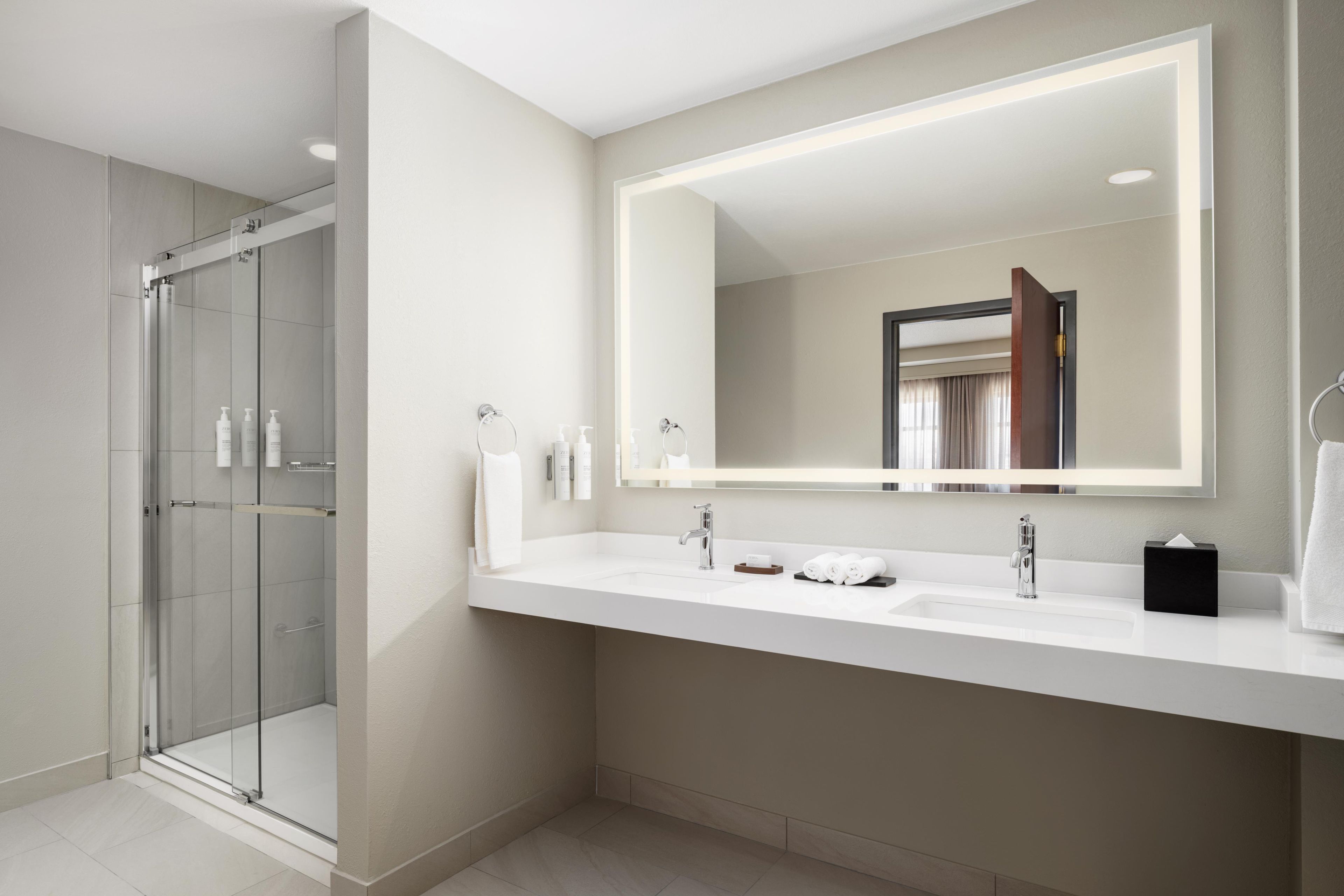 Un baño moderno con cabina de ducha de vidrio, un espejo grande y un tocador blanco con dos lavabos.