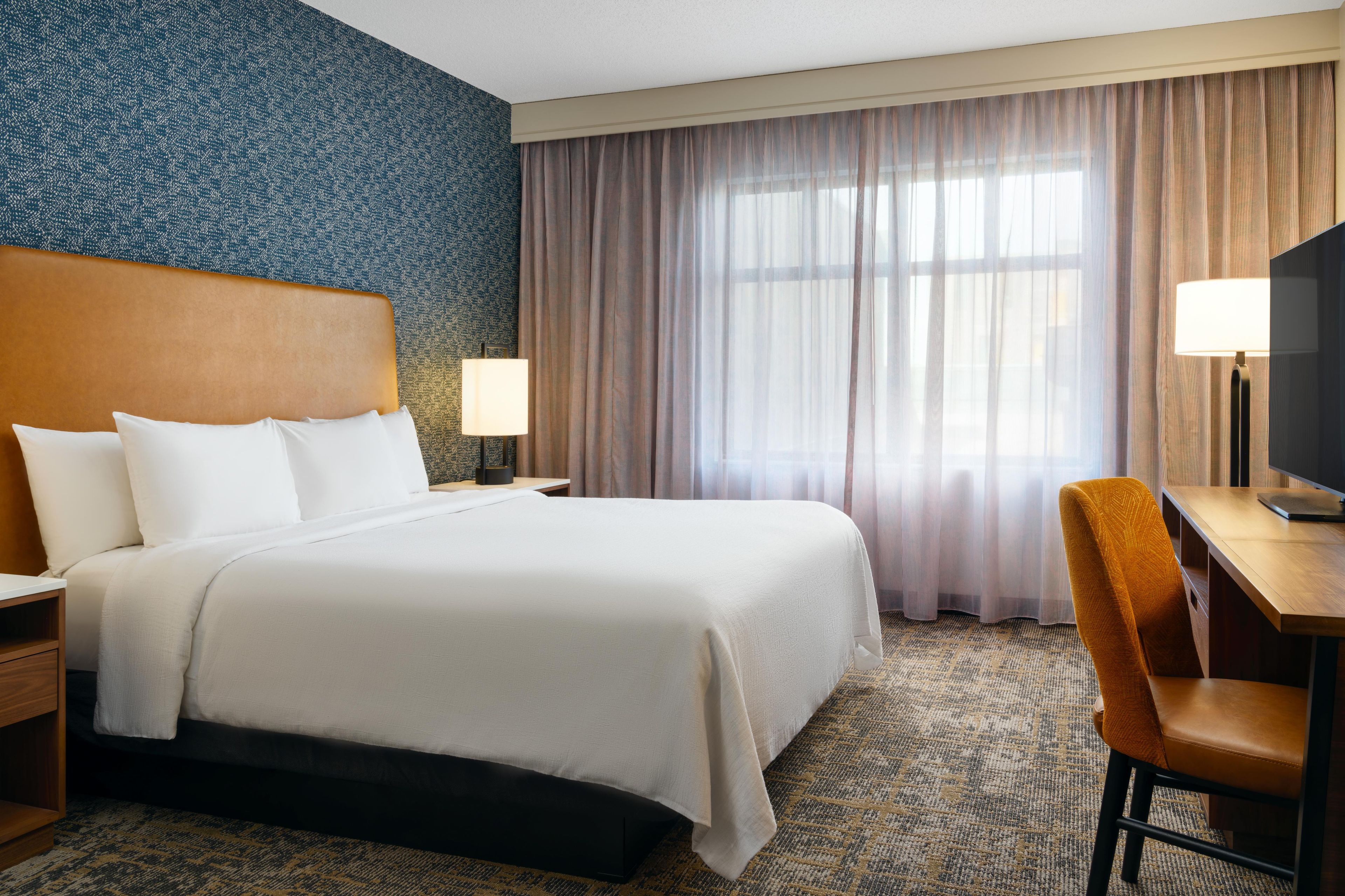 A hotel room with a large bed, a desk, a chair, a TV, and a window with curtains.