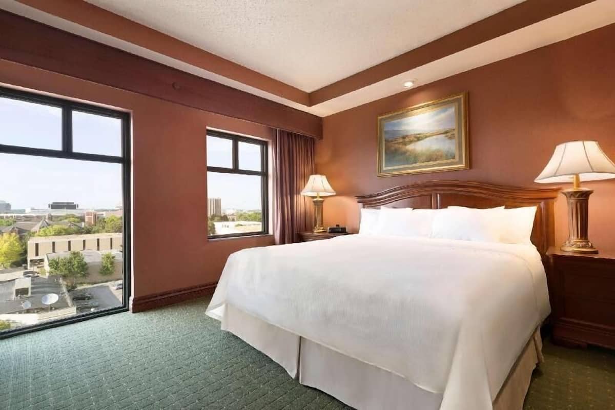 A hotel room with a large bed, two bedside lamps, a painting on the wall, and a window offering a city view.