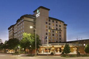 Un hotel de varios pisos con un letrero destacado que dice 'EMBASSY SUITES', un asta con la bandera estadounidense y una entrada bien iluminada.