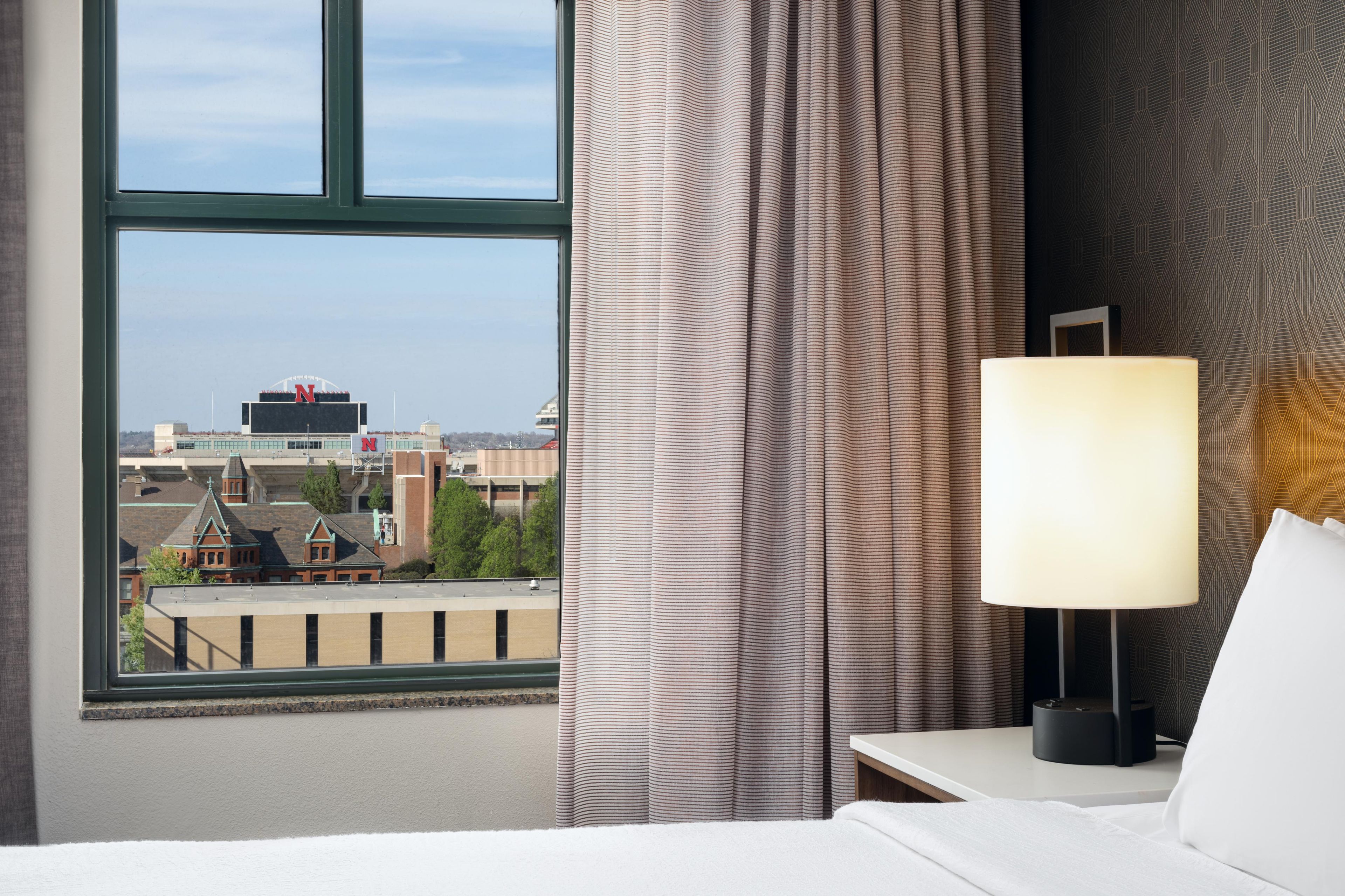 A hotel room with a bed, a nightstand, a lamp, and a view of a cityscape through a window with curtains.