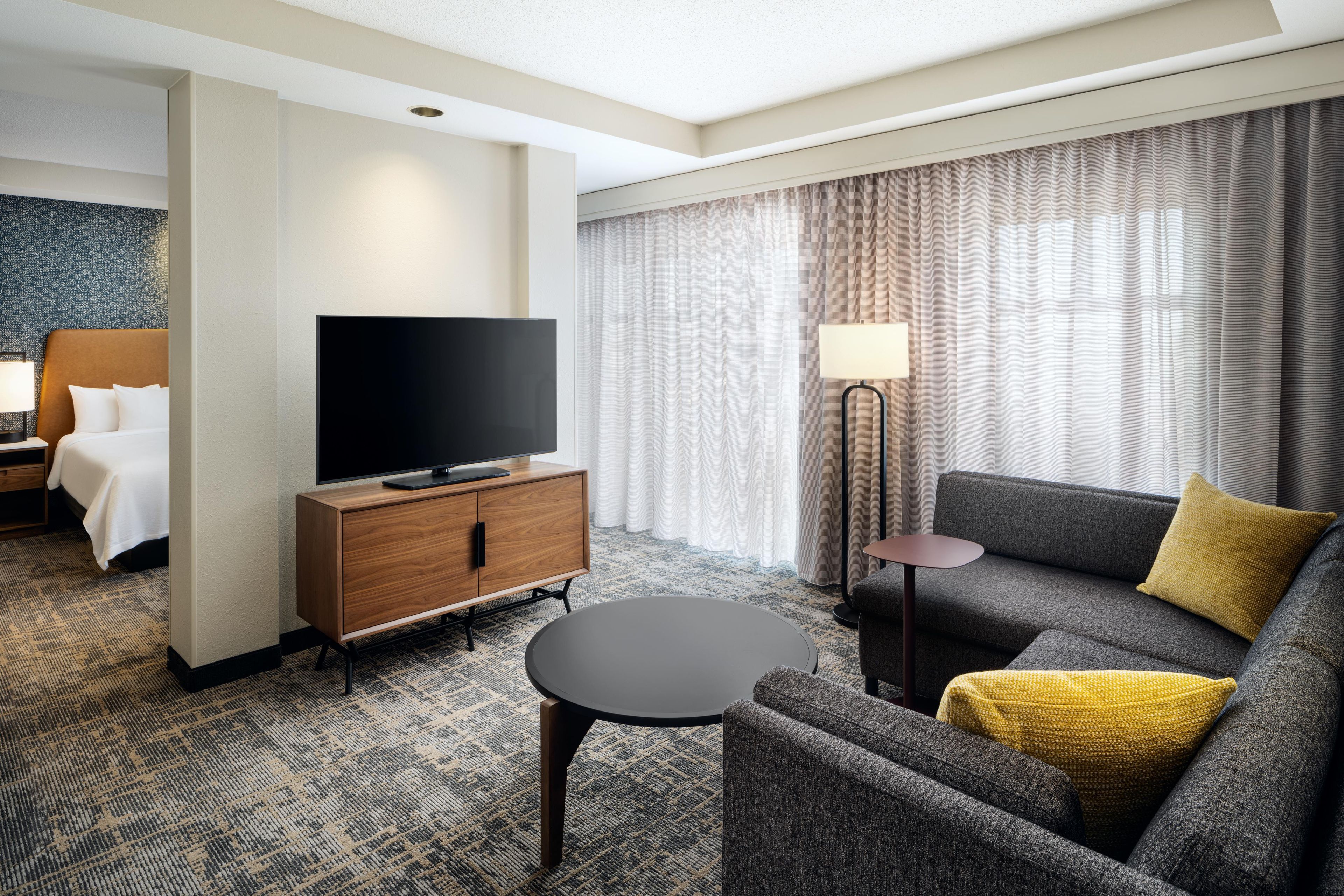 Una habitación de hotel con una televisión de pantalla plana sobre un mueble de madera, un sofá gris con un cojín amarillo y una mesa de centro redonda, además de una lámpara.