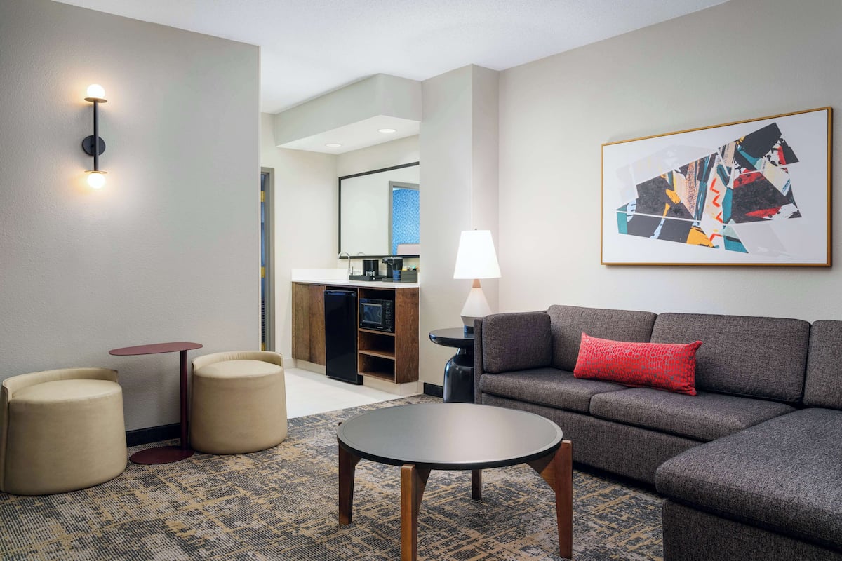 A modern hotel room with a grey sofa, two round ottomans, a small table, a lamp, and a framed abstract painting on the wall.