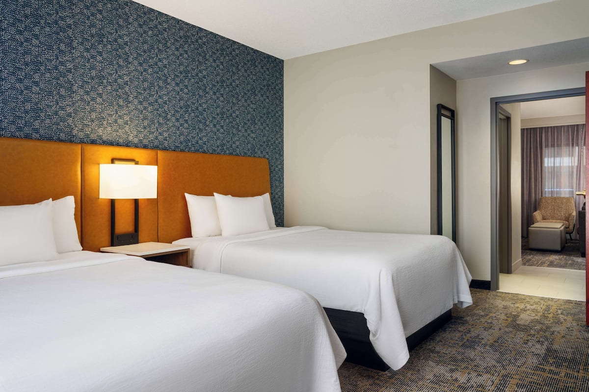 A hotel room with two beds, a headboard with a light, and a view of another room through an open door.