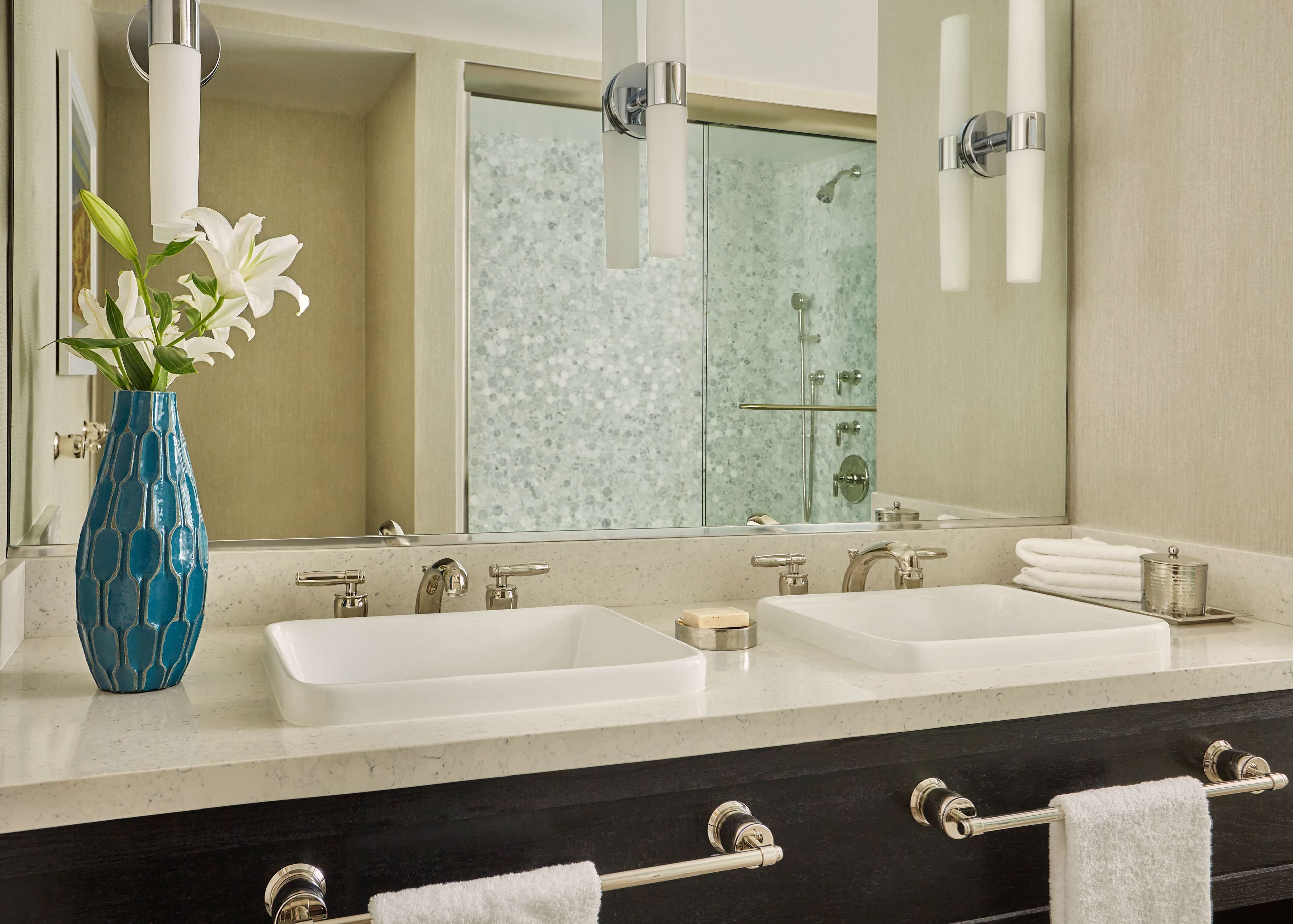 A bathroom with two sinks, a large mirror, a vase with flowers, and a shower area.