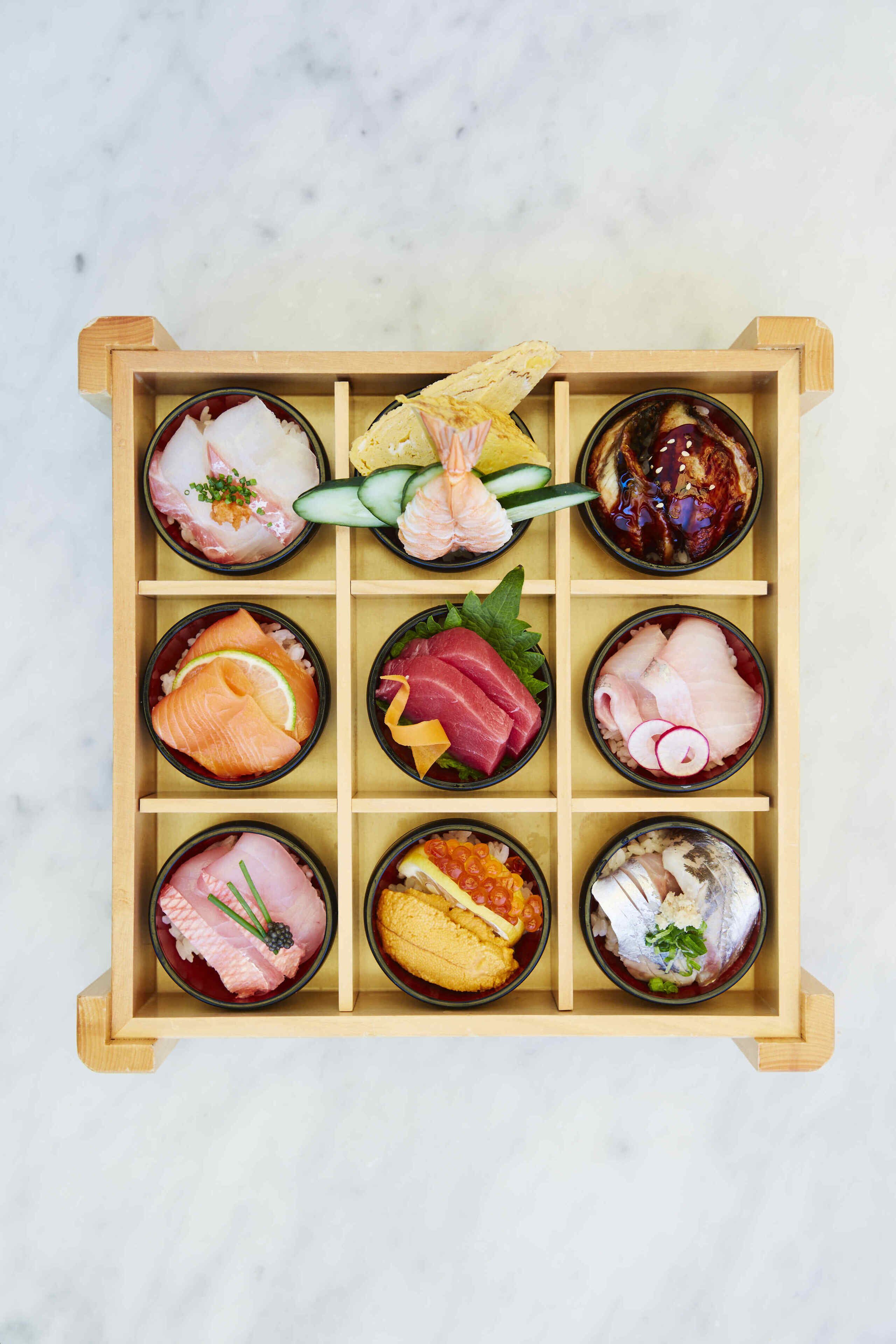 Una bandeja de madera con nueve recipientes que contienen diferentes tipos de sashimi y sushi.