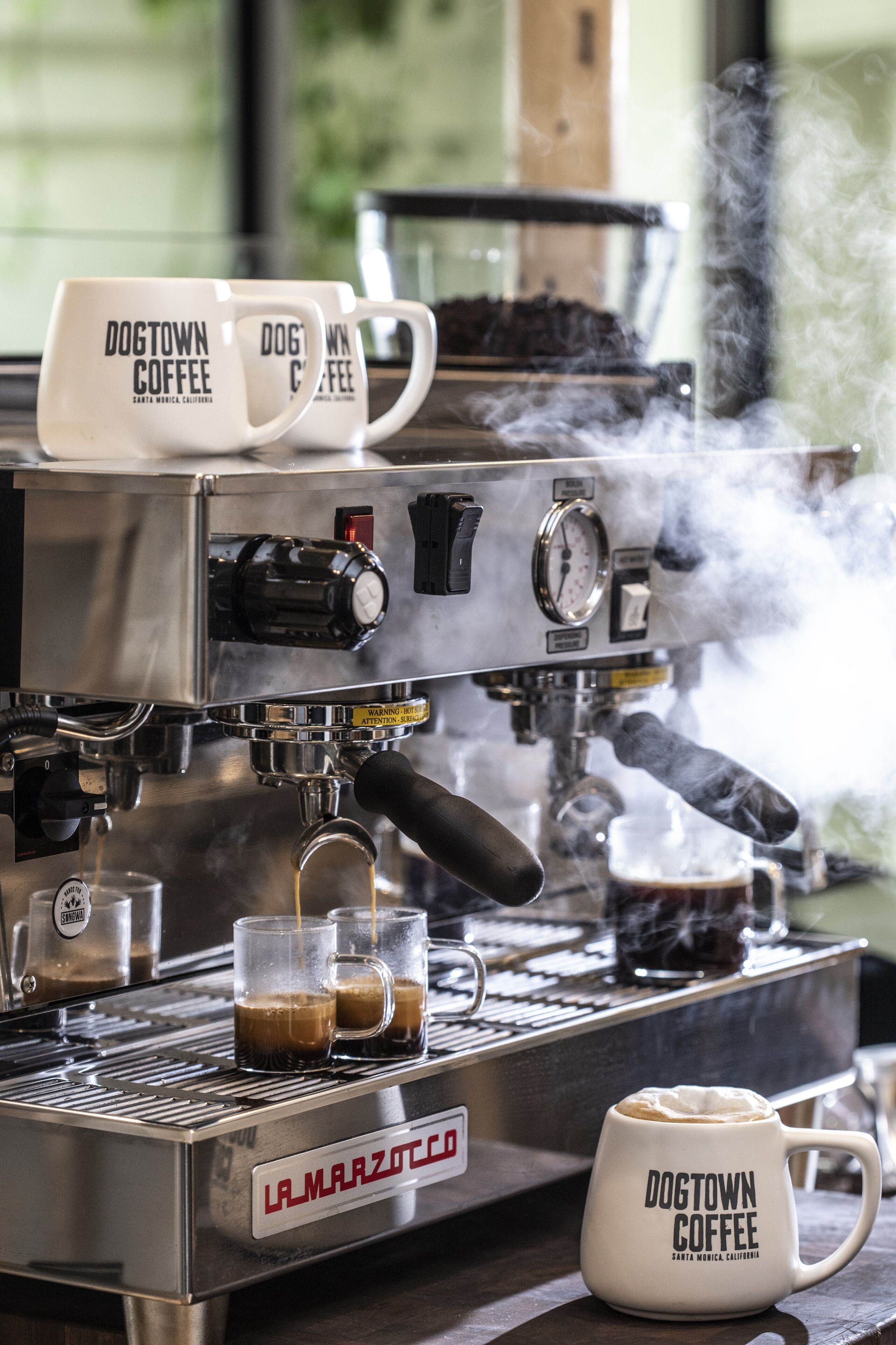 A coffee machine with steam, two mugs labeled "Dogtown Coffee," and two glasses of coffee on the counter.