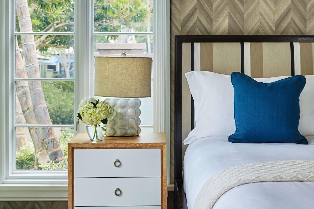 A bedroom with a bed, a nightstand with a lamp, and a window with a view of trees and a building.