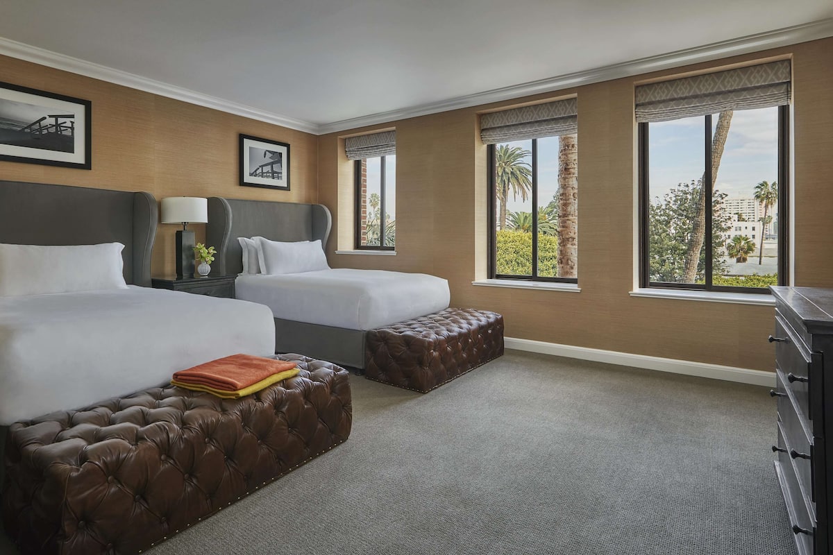 A hotel room with two beds, a large window with a view of palm trees, a brown leather ottoman, and a desk with a lamp.
