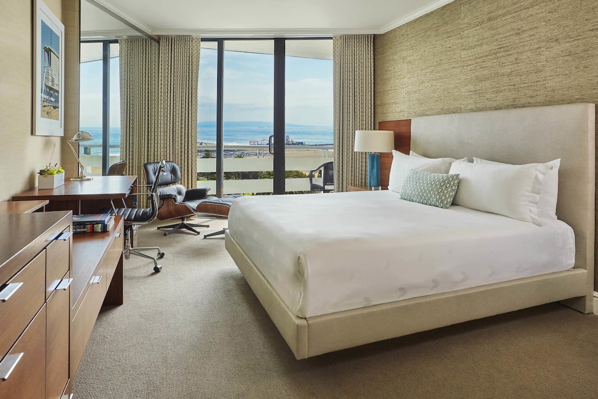 A hotel room with a large bed, a desk with a computer, a chair, and a view of the beach.