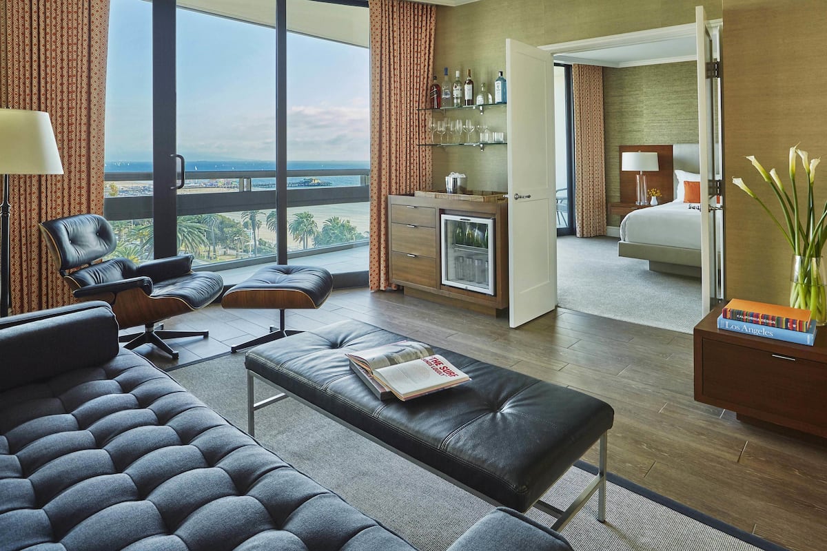 A modern hotel room with a large window, a seating area with a sofa and ottoman, a minibar, and a view of the cityscape.