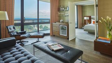 A modern hotel room with a large window, a seating area with a sofa and ottoman, a minibar, and a view of the cityscape.