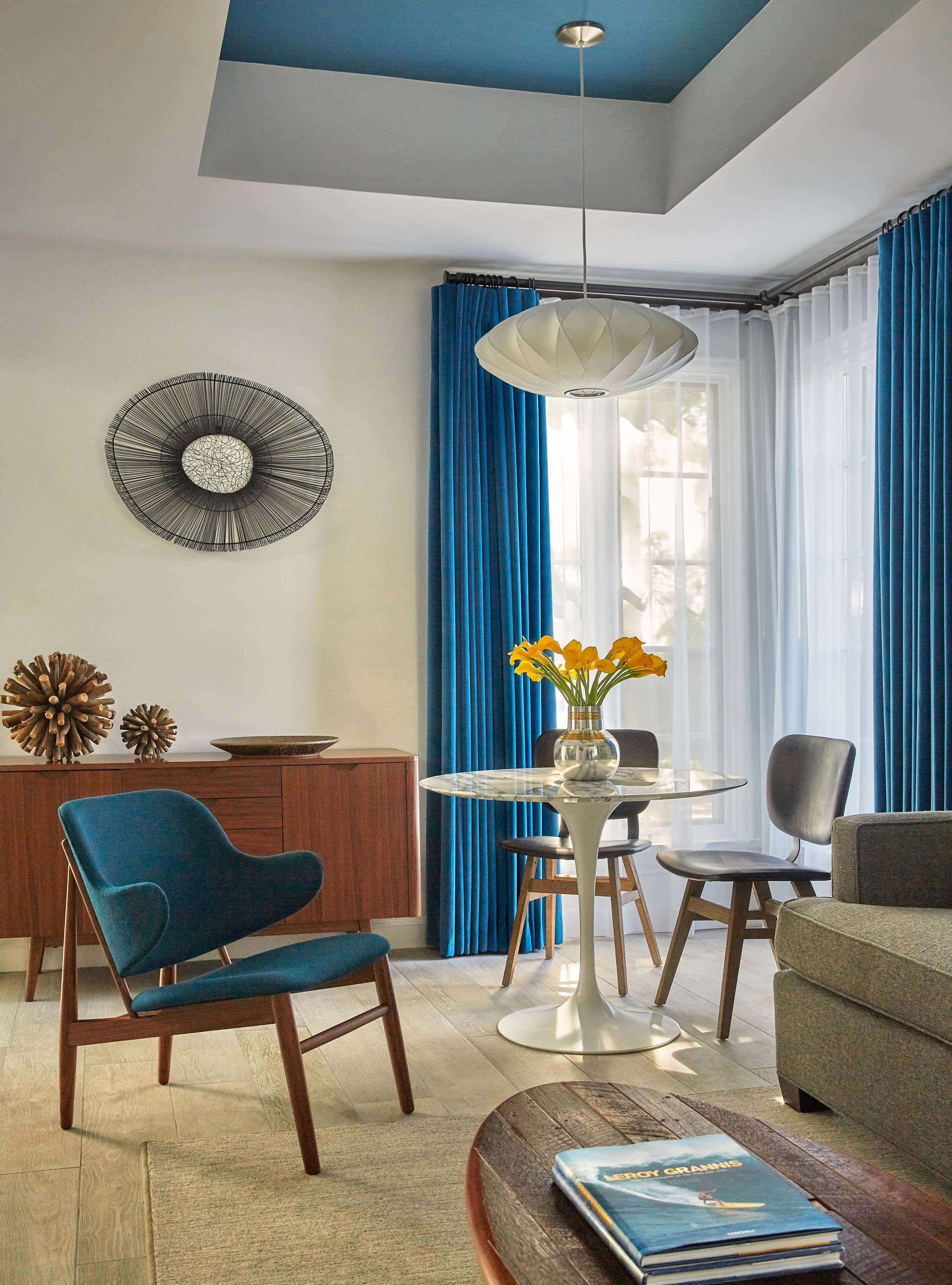 A modern living room with a blue chair, a round table with a vase of flowers, and a dining area with a wooden cabinet and blue curtains.