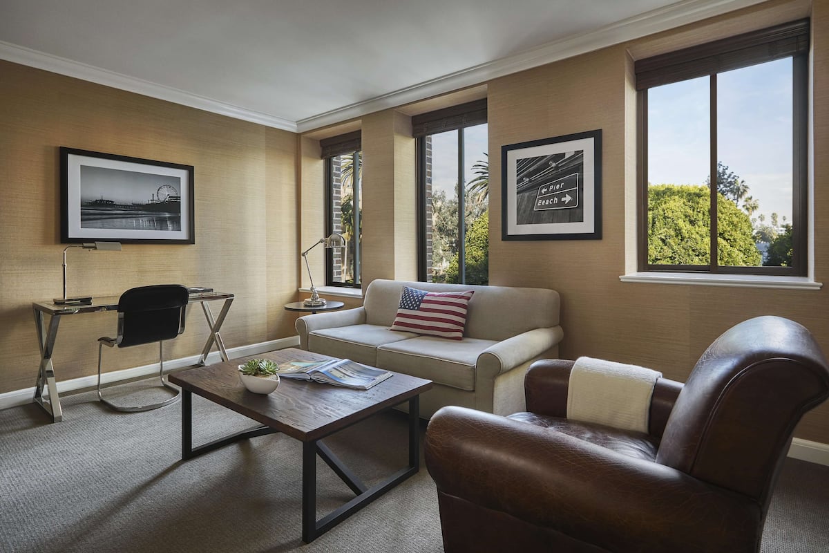 A living room with a beige sofa, a brown leather armchair, a coffee table with a book, a desk with a chair, and two framed pictures on the wall.