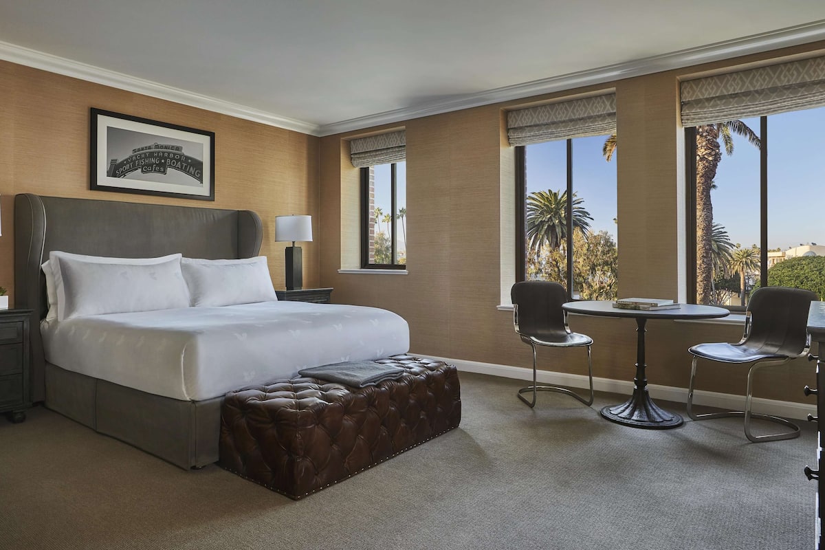 A hotel room with a large bed, two bedside lamps, a small table with chairs, and a view of palm trees through the windows.
