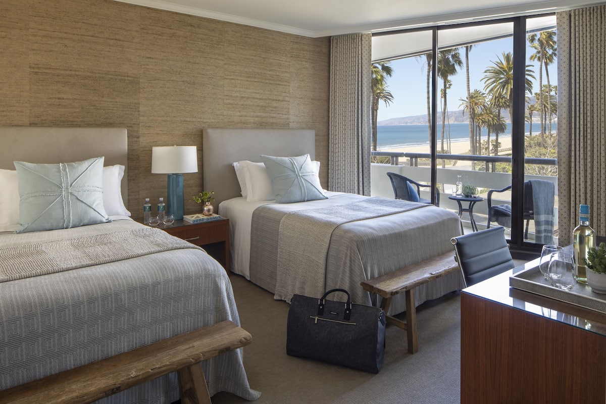 A hotel room with two beds, a desk, a bench, and a view of the beach and palm trees.