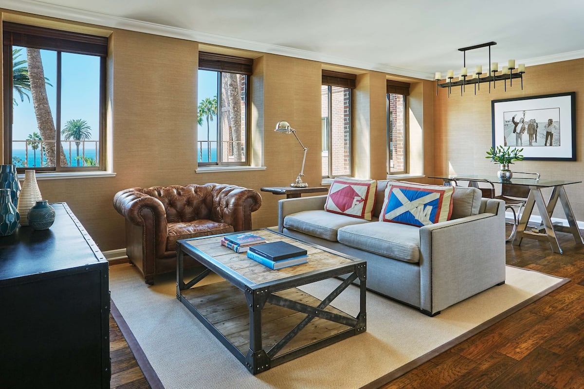A living room with a leather armchair, a gray sofa with colorful pillows, a coffee table, and a dining area with a metal table and chairs.