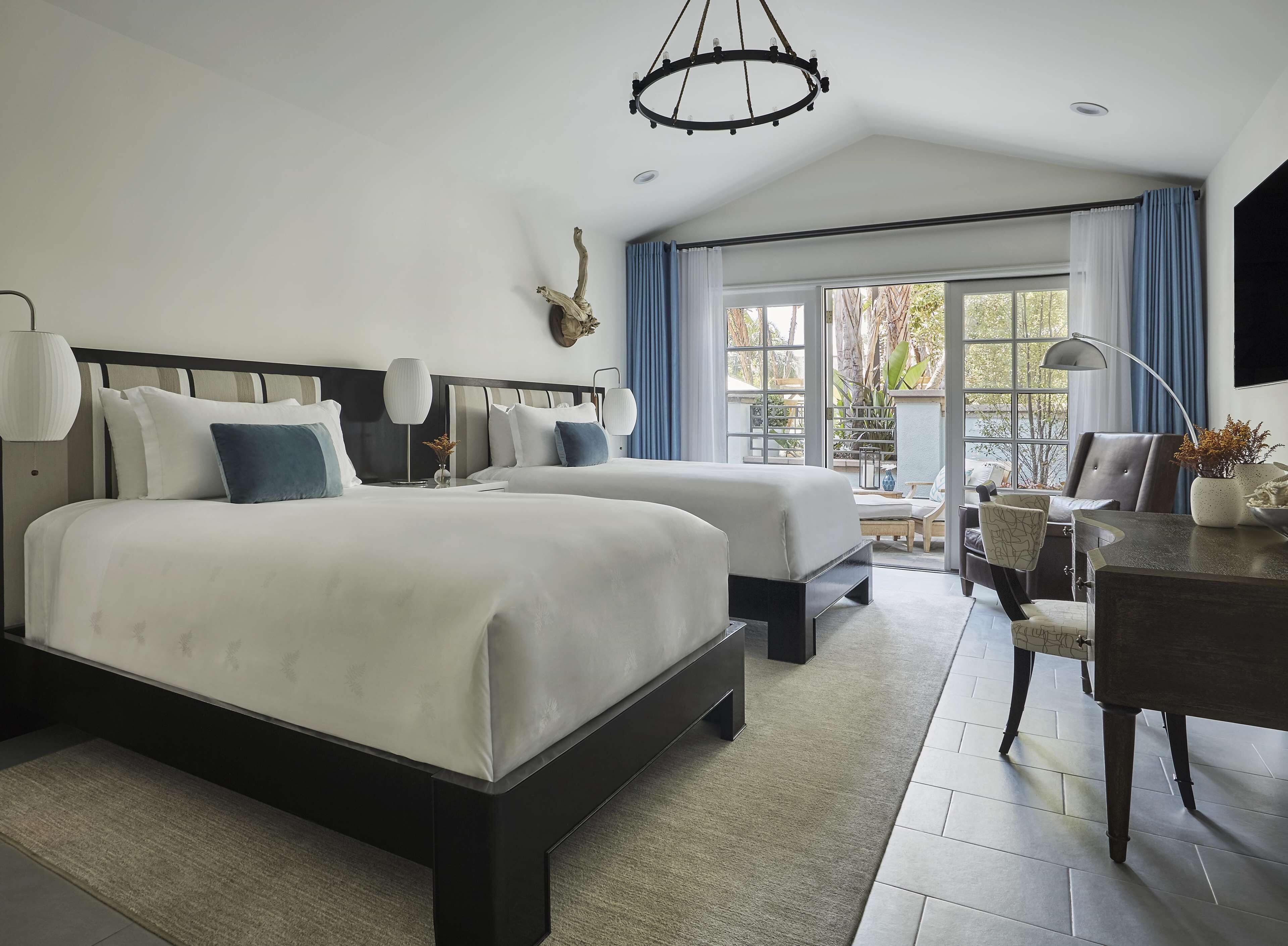 A hotel room with two beds, a desk, a chair, and a view of the outdoors.