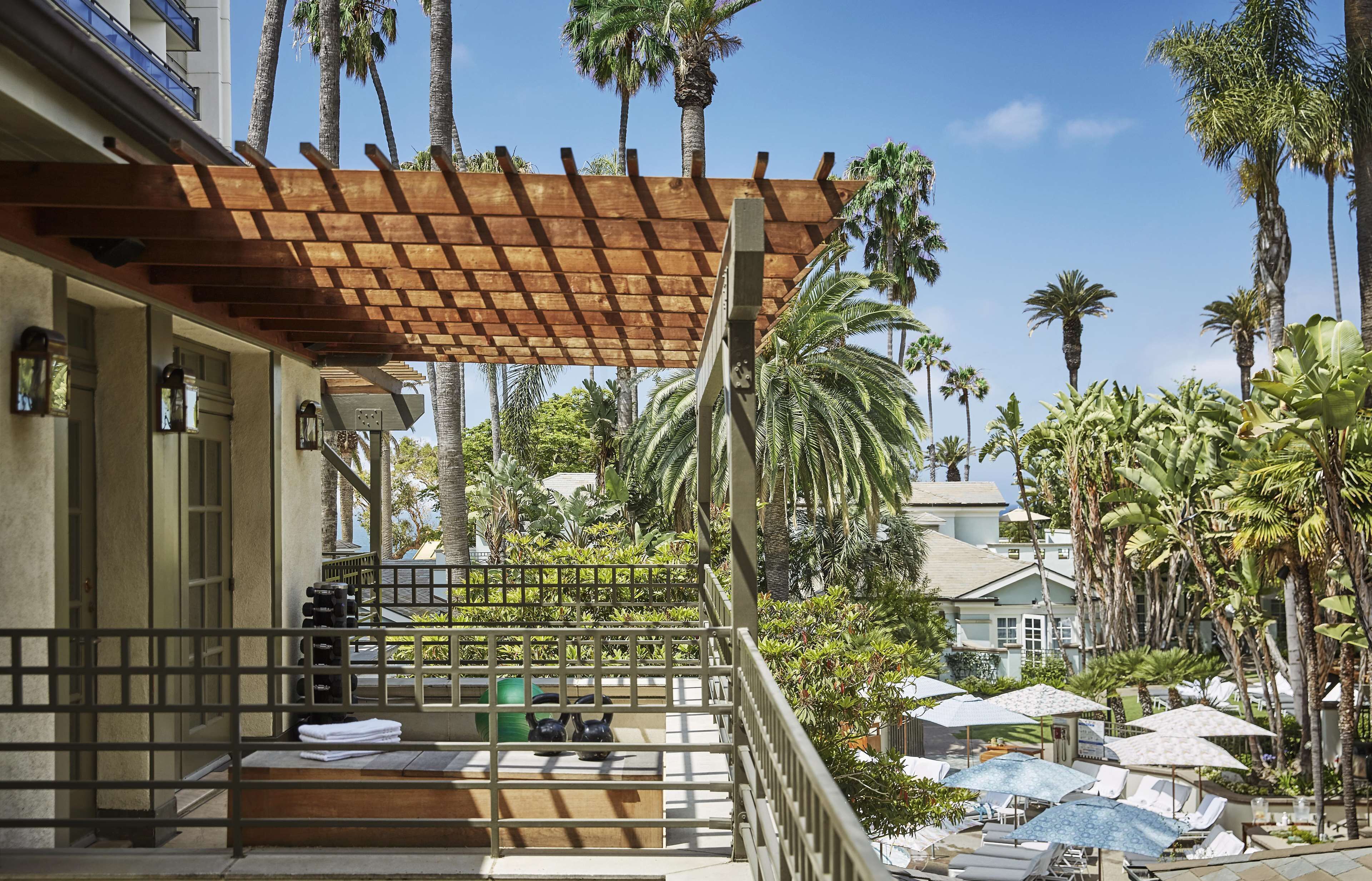 Varanda de hotel com pérgola de madeira, palmeiras e área de estar externa com guarda-sóis.