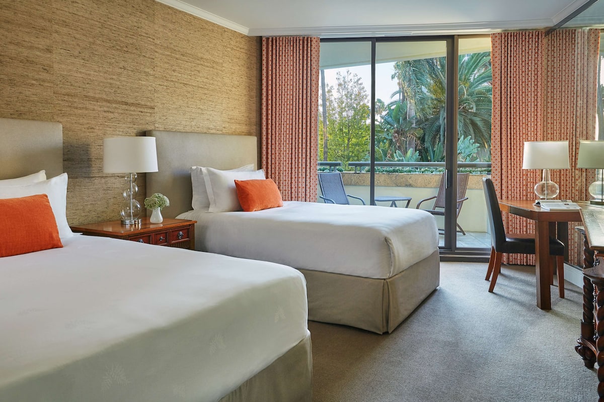 A hotel room with two beds, a desk, a chair, and a view of the outdoors.