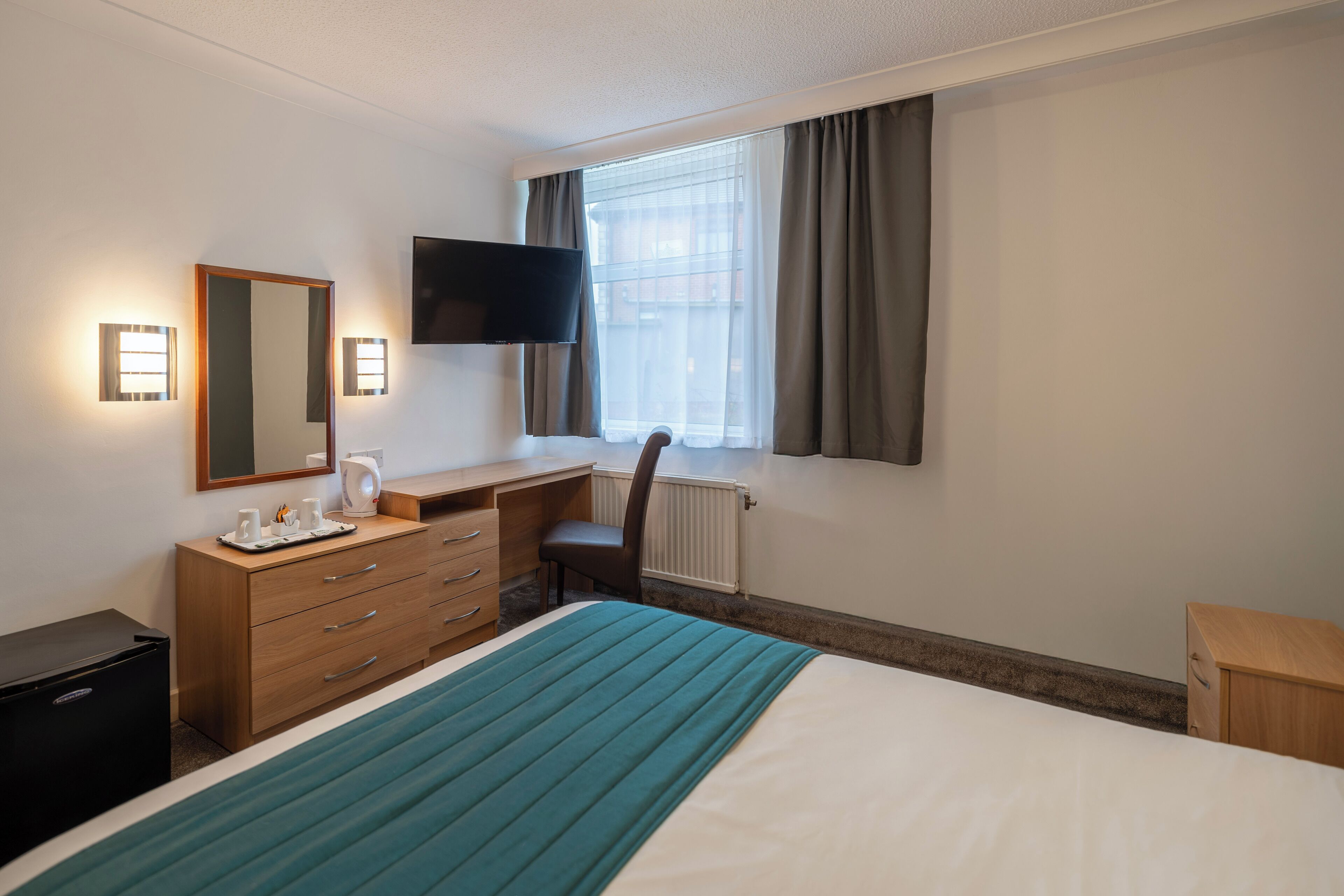 Habitación de hotel con cama, escritorio, silla, televisor y ventana con cortinas.
