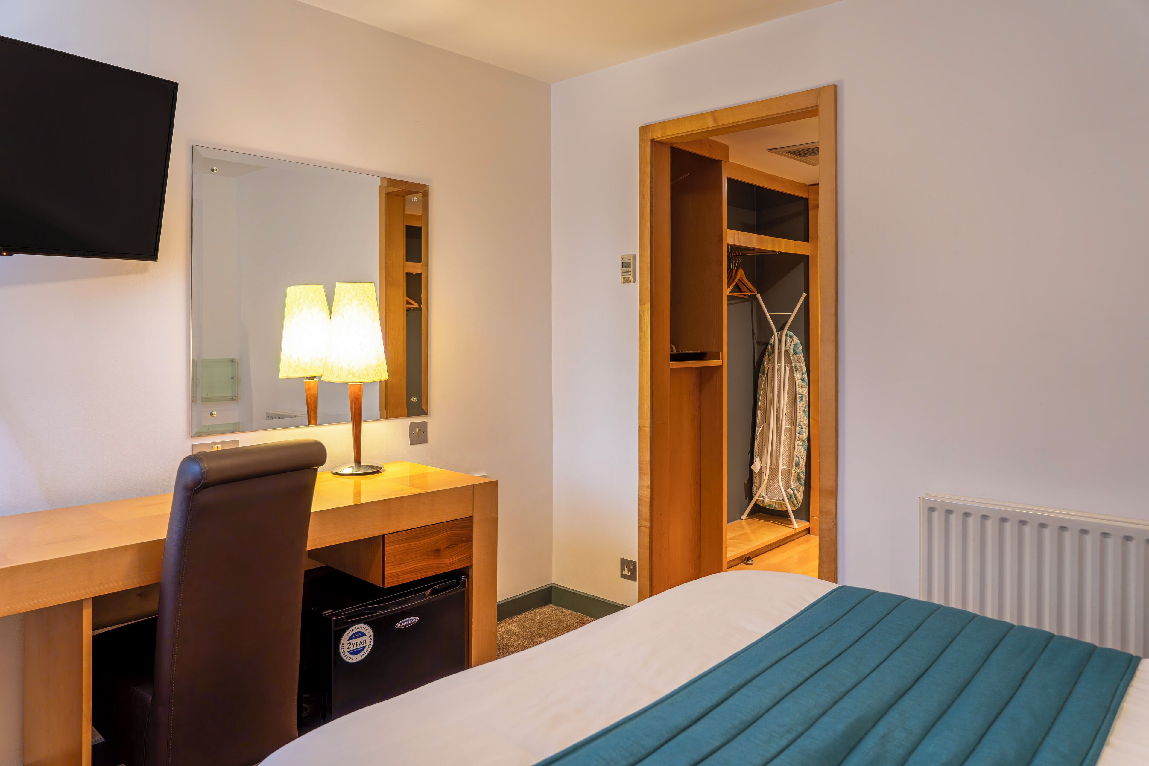 A hotel room with a bed, a desk with a chair, a TV mounted on the wall, and a wooden wardrobe.