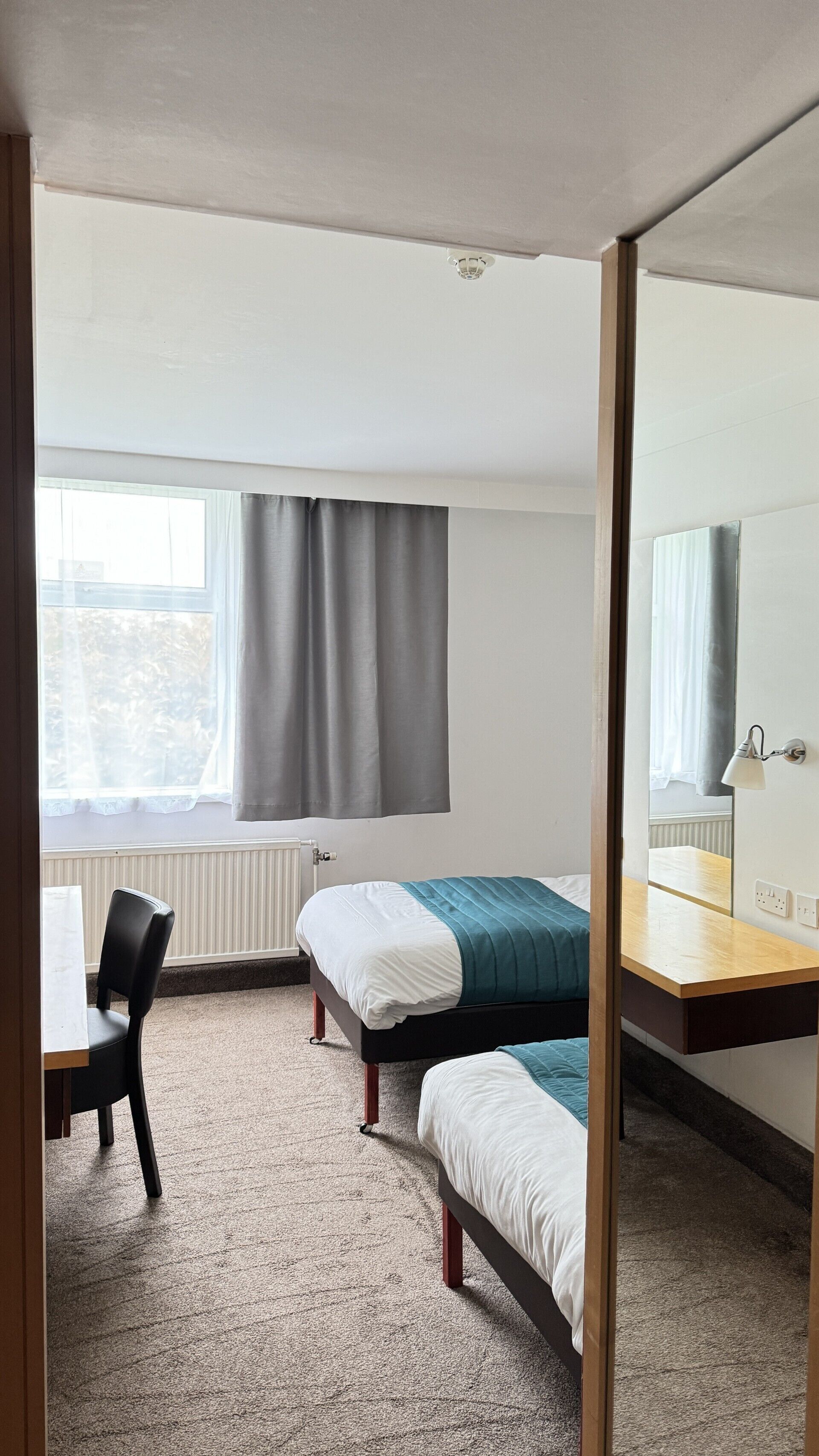A small, tidy room with two single beds, a desk, a chair, and a window with curtains.