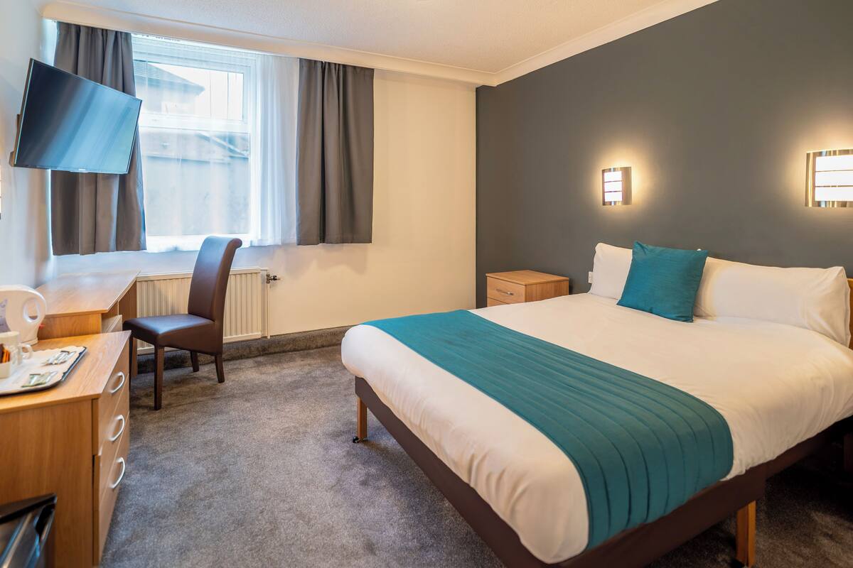 Habitación de hotel con cama, escritorio, silla, televisor y ventana con cortinas.