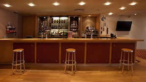 Una zona de bar con mostrador de madera, tres taburetes y estanterías llenas de botellas.