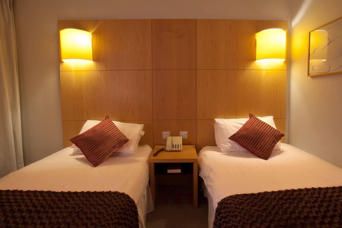 Habitación de hotel con dos camas, un cabecero de madera, lámparas de noche, una mesita de noche con un teléfono y un cuadro en la pared.