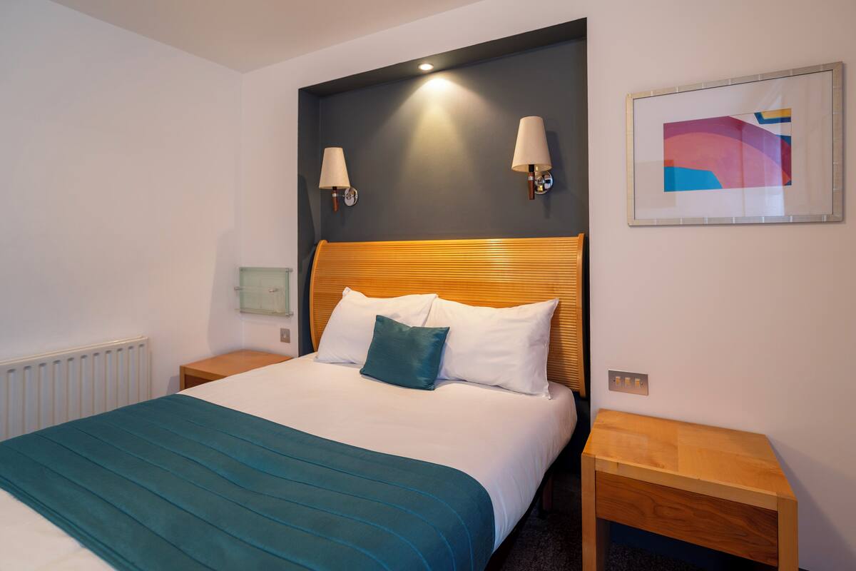Una cama bien hecha con una colcha color turquesa y almohadas blancas, dos lámparas de pared, un cuadro abstracto enmarcado y una mesita de noche de madera.