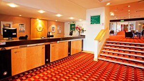 Zona de recepción de un hotel con mostrador de registro, una escalera e indicaciones de servicios como restaurante y bar.