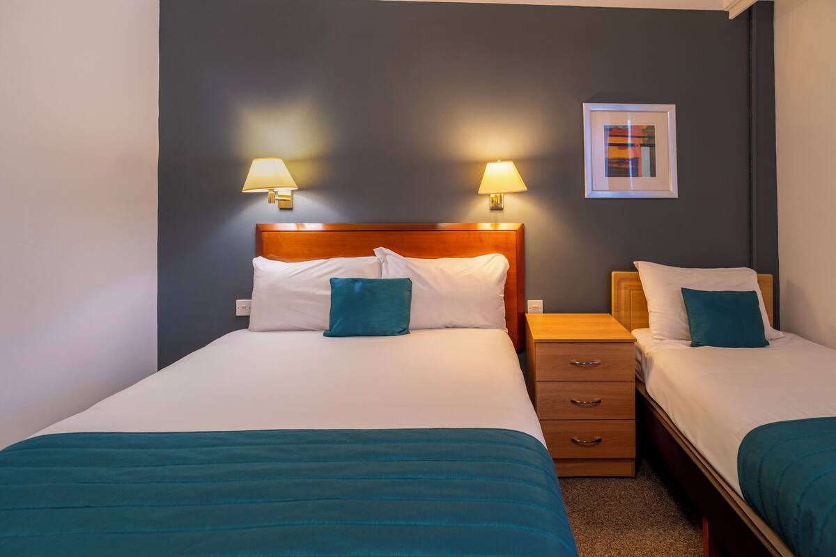 Habitación de hotel con dos camas, cada una con cabecero de madera, ropa de cama blanca y almohadones color aguamarina. Hay dos mesitas de noche con lámparas, y un cuadro enmarcado en la pared.