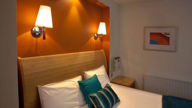 Habitación de hotel con cabecera de madera, dos lámparas de noche, una obra de arte enmarcada en la pared y una cama con almohadas blancas y color turquesa.