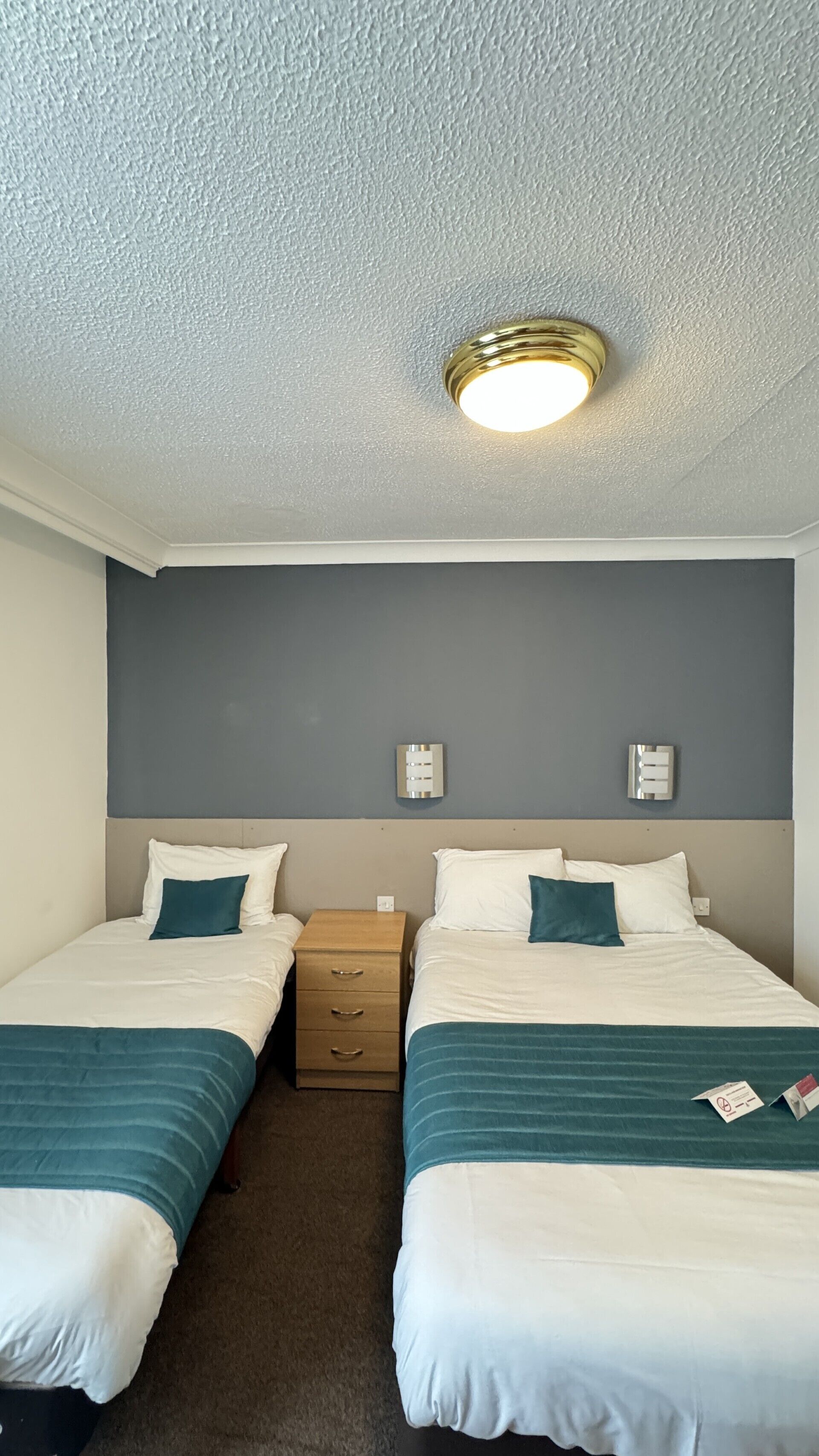 A hotel room with two beds, a nightstand, a wall-mounted light fixture, and a thermostat.