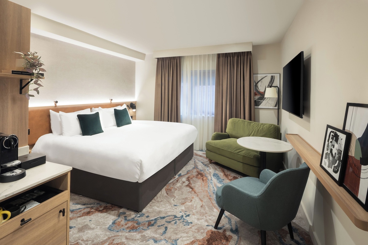 Habitación de hotel con una cama grande, un escritorio con cafetera, una silla, una mesita y un televisor montado en la pared.